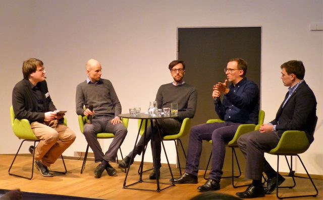 Fünf Männer sitzen auf Stühlen auf einem Podium und diskutieren