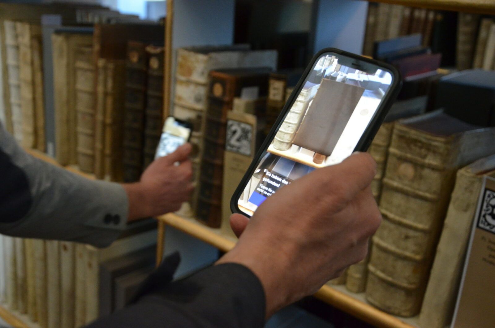 Zwei Hände mit Smartphone, auf dem Display rechts ist ein Buch als 3D-Objekt zu sehen, auf dem Display links ist ein AR-Marker (unscharf) zu erkennen