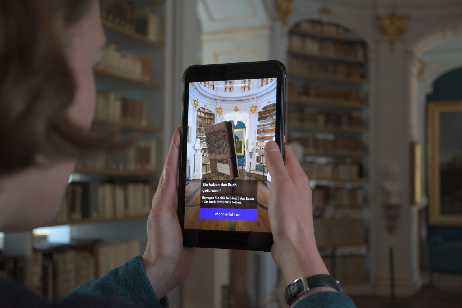 Blick über die Schulter einer Nutzerin im Rokokosaal, sie hält ein Tablet, auf dem ein in AR animiertes 3D-Buch zu sehen ist.