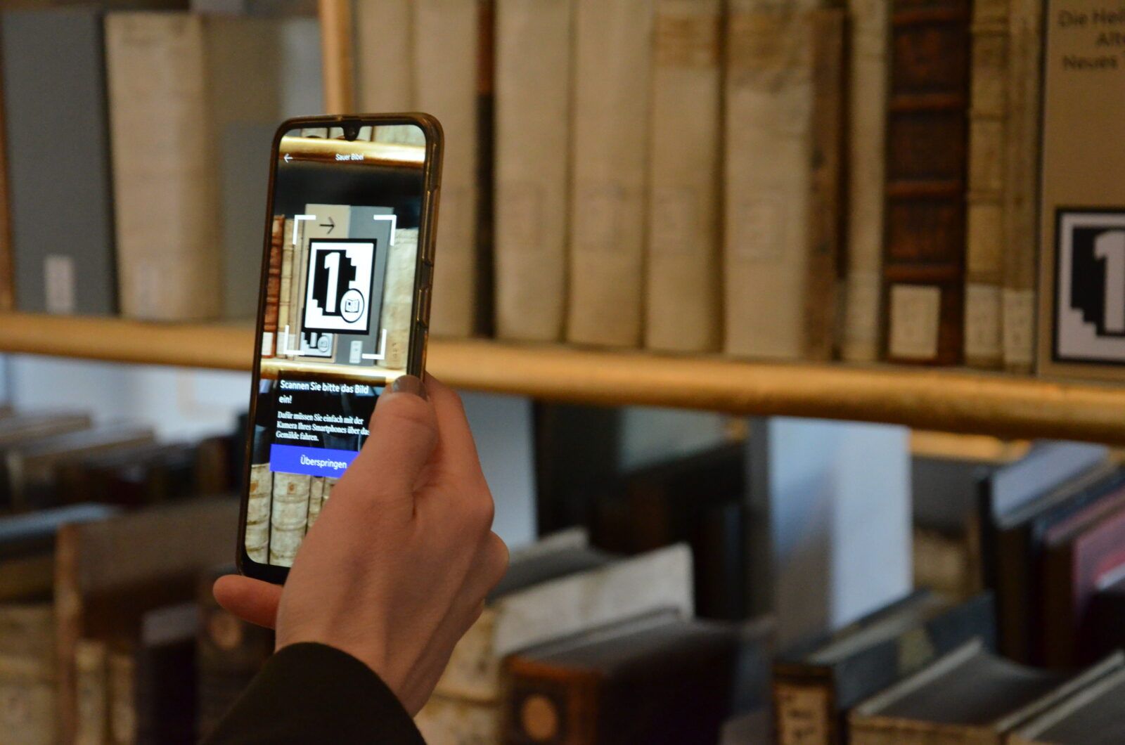 Hand mit Smartphone vor Bücherregal im Rokokosaal, auf dem Screen ist ein AR-Marker eingeblendet