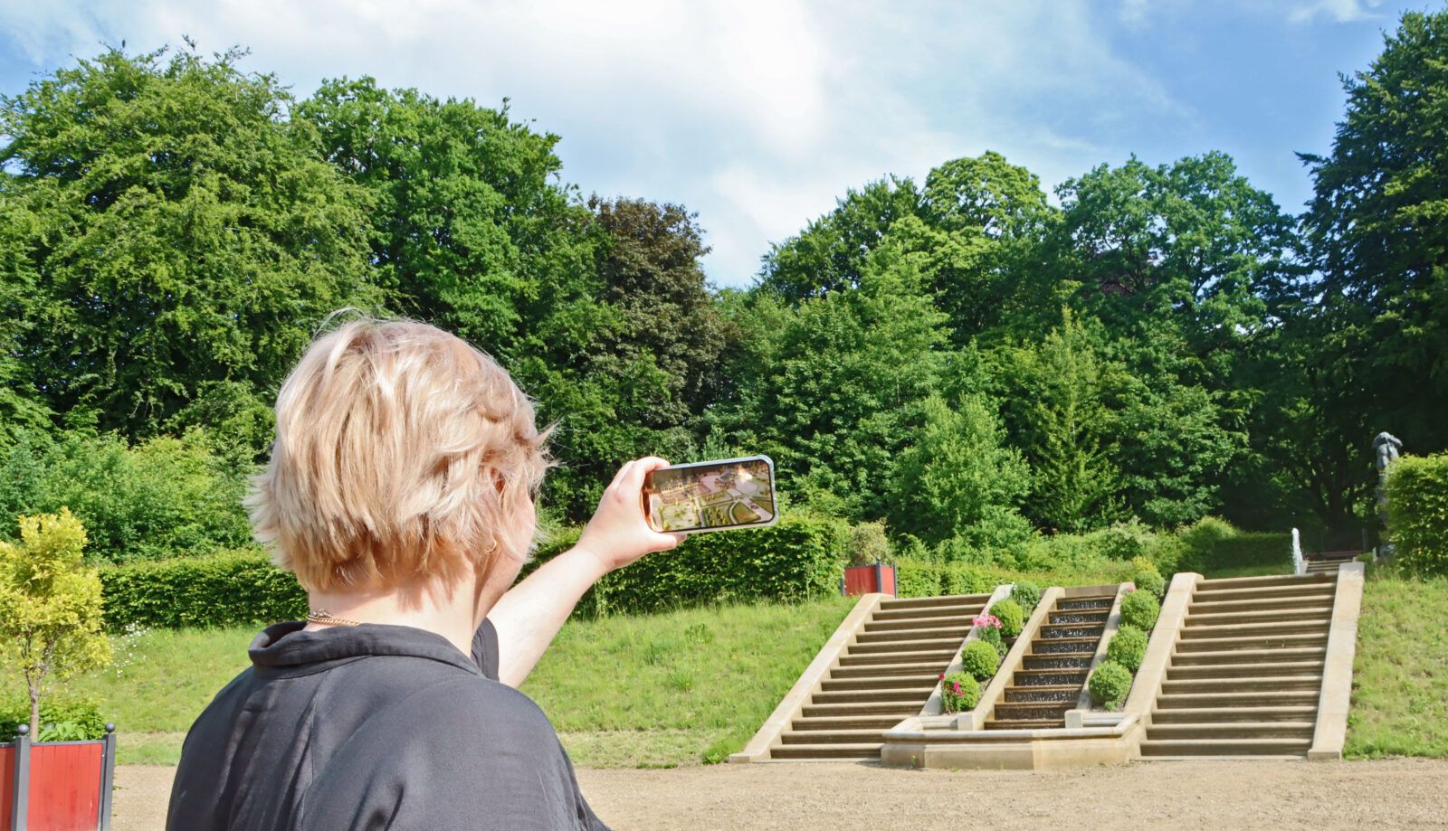 Das Bild zeigt die Anwendung einer AR-Szene im heutigen Brockgarten. Eine Person hält ein Smartphone hoch, auf dem ein animiertes 3D-Modell des historischen Barockgartens aus dem 17. Jahrhundert dargestellt ist. Foto: Stiftung Schleswig-Holsteinische Landesmuseen Schloss Gottorf, CC BY 4.0