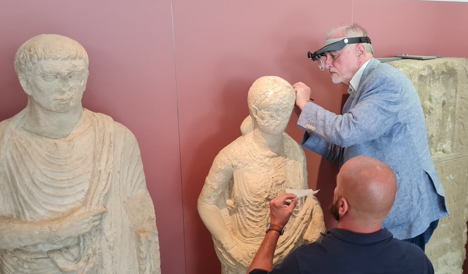 Ein älterer Mann mit einer Stirnlupe und einem Skalpell arbeitet konzentriert am Kopf einer lebensgroßen Grabfigur. Ein zweiter Mann hilft ihm.