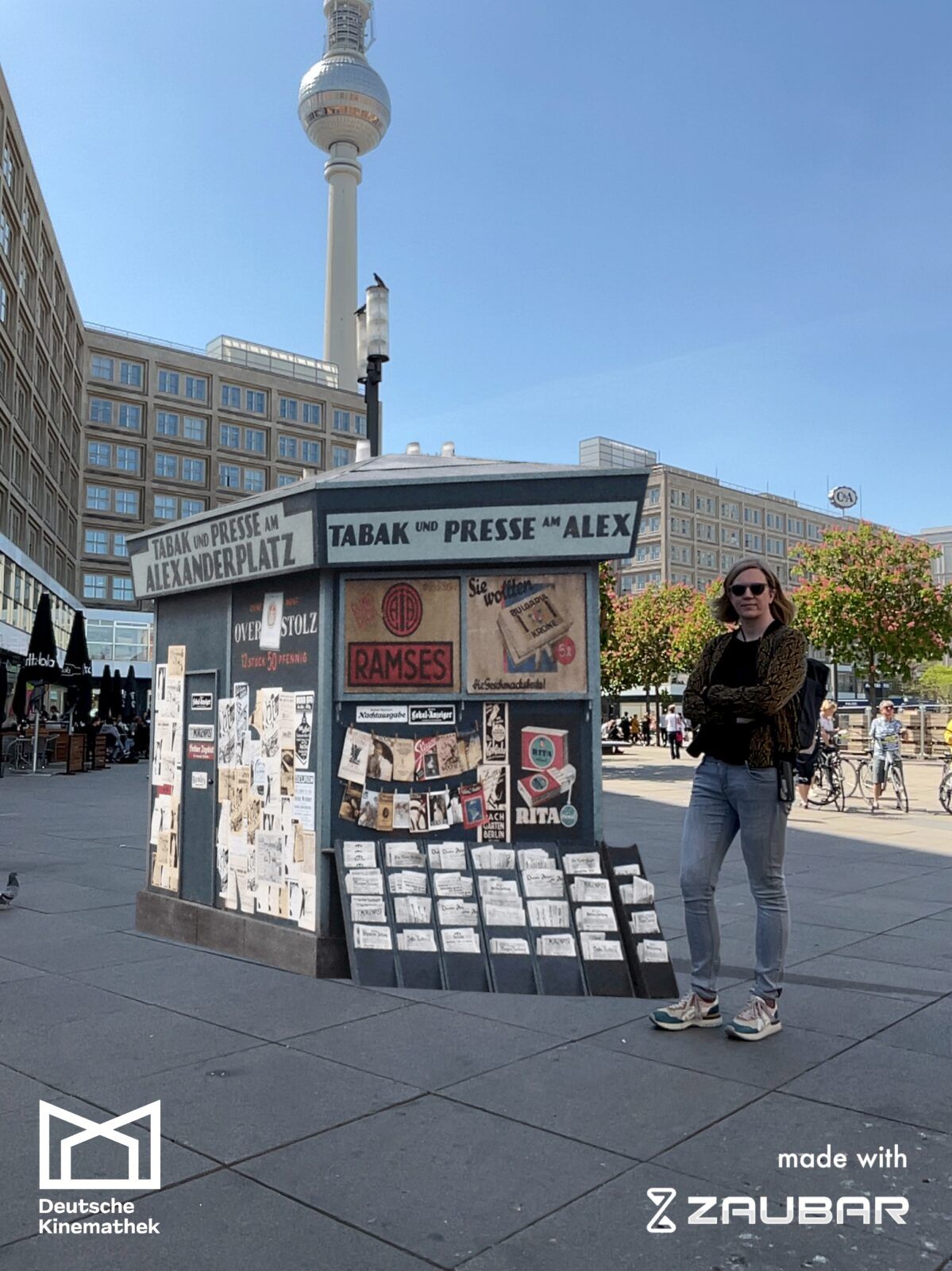 Das Foto zeigt einen Zeitungskiosk aus der 1920er-Jahren, der beim Dreh der Serie „Babylon Berlin“ verwendet wurde. Dieser steht hier virtuell auf dem Alexanderplatz in Berlin. Daneben steht eine Frau. Dies ist möglich mit Augmented Reality.