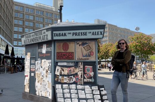 Das Foto zeigt einen Zeitungskiosk aus der 1920er-Jahren, der beim Dreh der Serie „Babylon Berlin“ verwendet wurde. Dieser steht hier virtuell auf dem Alexanderplatz in Berlin. Daneben steht eine Frau. Dies ist möglich mit Augmented Reality.