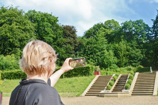 Das Bild zeigt die Anwendung einer AR-Szene im heutigen Brockgarten. Eine Person hält ein Smartphone hoch, auf dem ein animiertes 3D-Modell des historischen Barockgartens aus dem 17. Jahrhundert dargestellt ist
