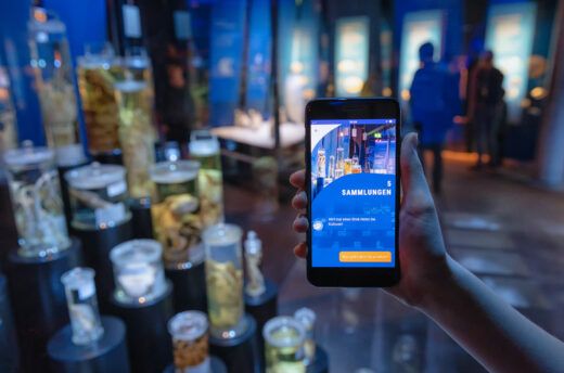 Das Bild zeigt im Vordergrund eine Smartphone mit der Museumsapp 
