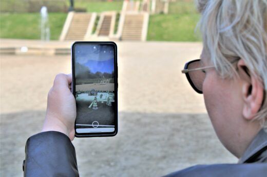 Das Foto zeigt eine Frau, die in einem barocken Park ihr Handy mit einer Augmented Reality Anwendung hält.