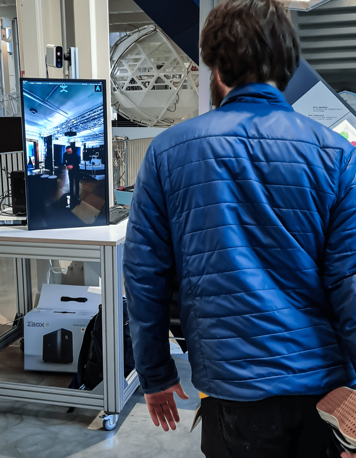 Auf dem Bild steht ein Mann mit dem Rücken zum Bild und schaut auf ein übergroßes Tablet, dass einen Mann in einem Museumsraum zeigt.
