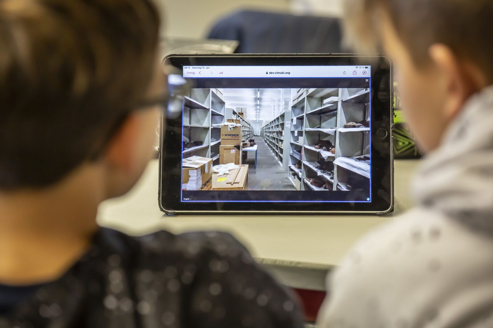 Zwei Kinder schauen auf ein Tablet, das die Depot-Räume eines Museums zeigt.