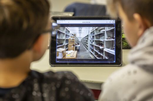 Zwei Kinder schauen auf ein Tablet, das die Depot-Räume eines Museums zeigt.