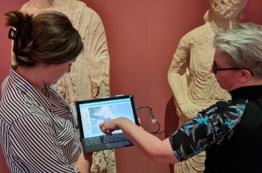 Vor zwei, zum Teil stark verwitterten römischen Stauten stehen zwei Frauen. Die eine hält einen Laptop mit Forschungsergebnissen, während die andere auf den Bildschirm zeigt.