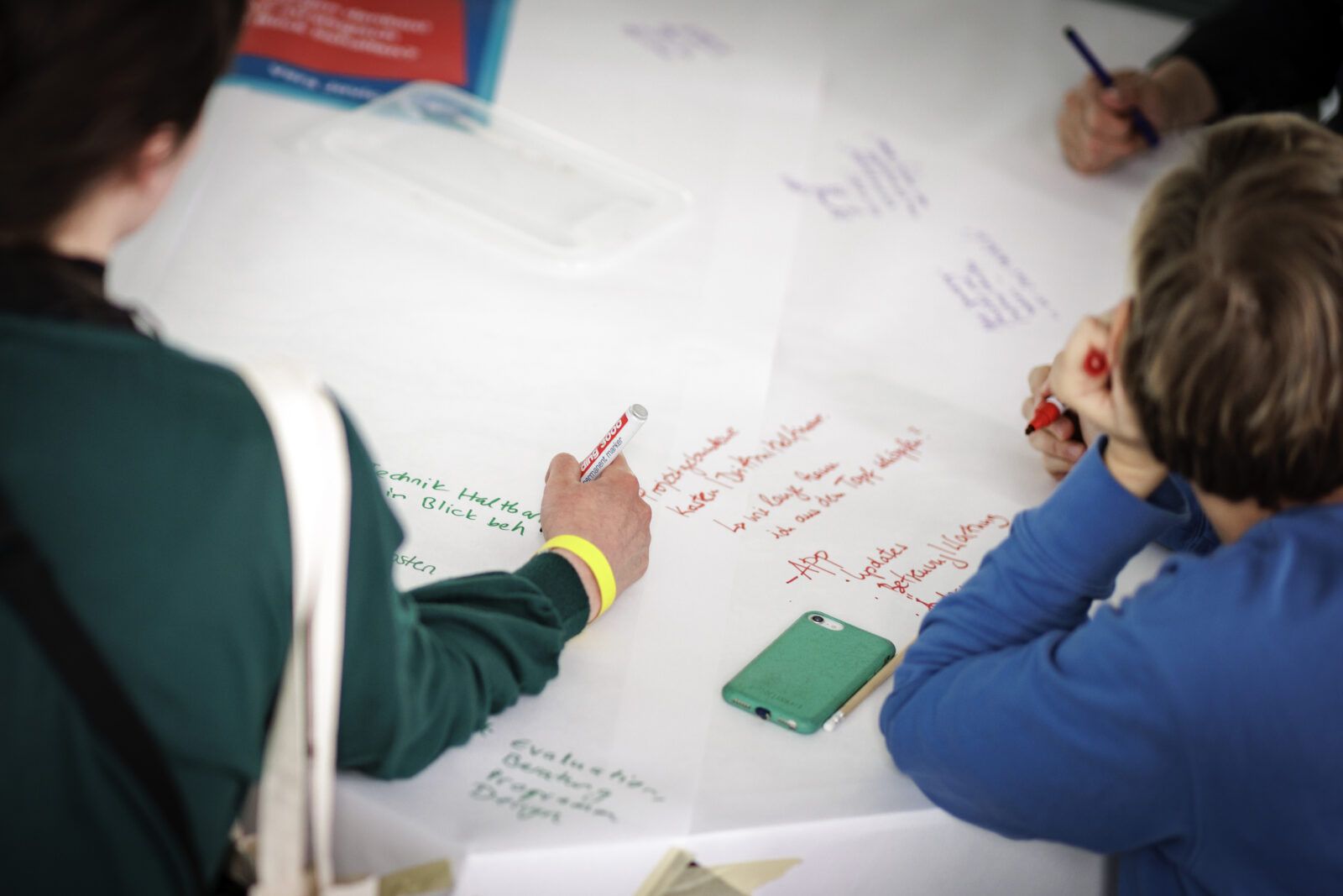 Das Bild zeigt Personen, die während eines World-Café Formates um einen Tisch Brainstormen und die Ergebnisse niederschreiben.
