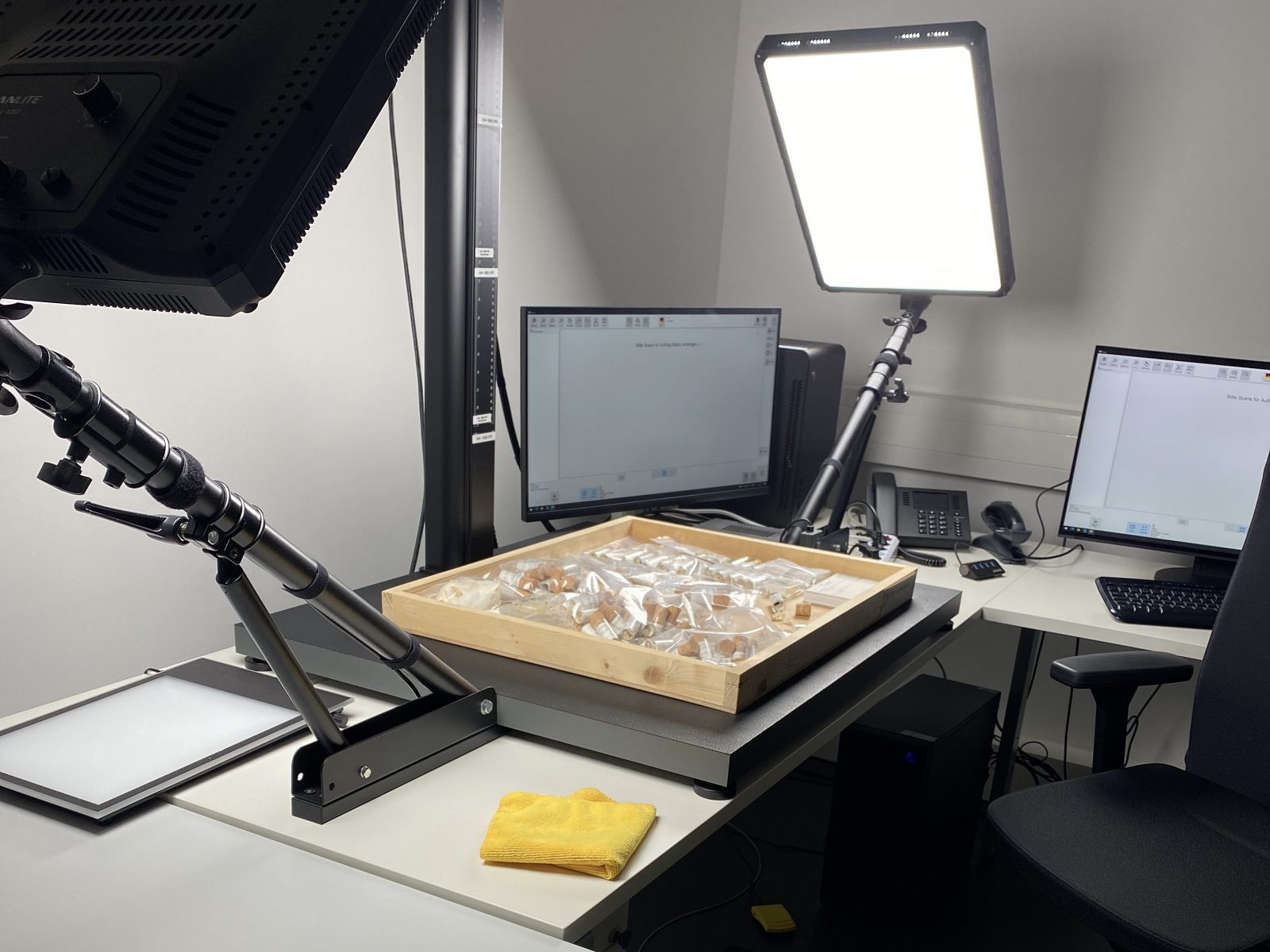 Die Schublade mit Kleinsäugerschädeln liegt zum Fotografieren bereit auf einer Arbeitsplatte unterhalb der Kamera, während sie von zwei flachen Lampen angeleuchtet wird und im Hintergund sind zwei Computerbildschirme.