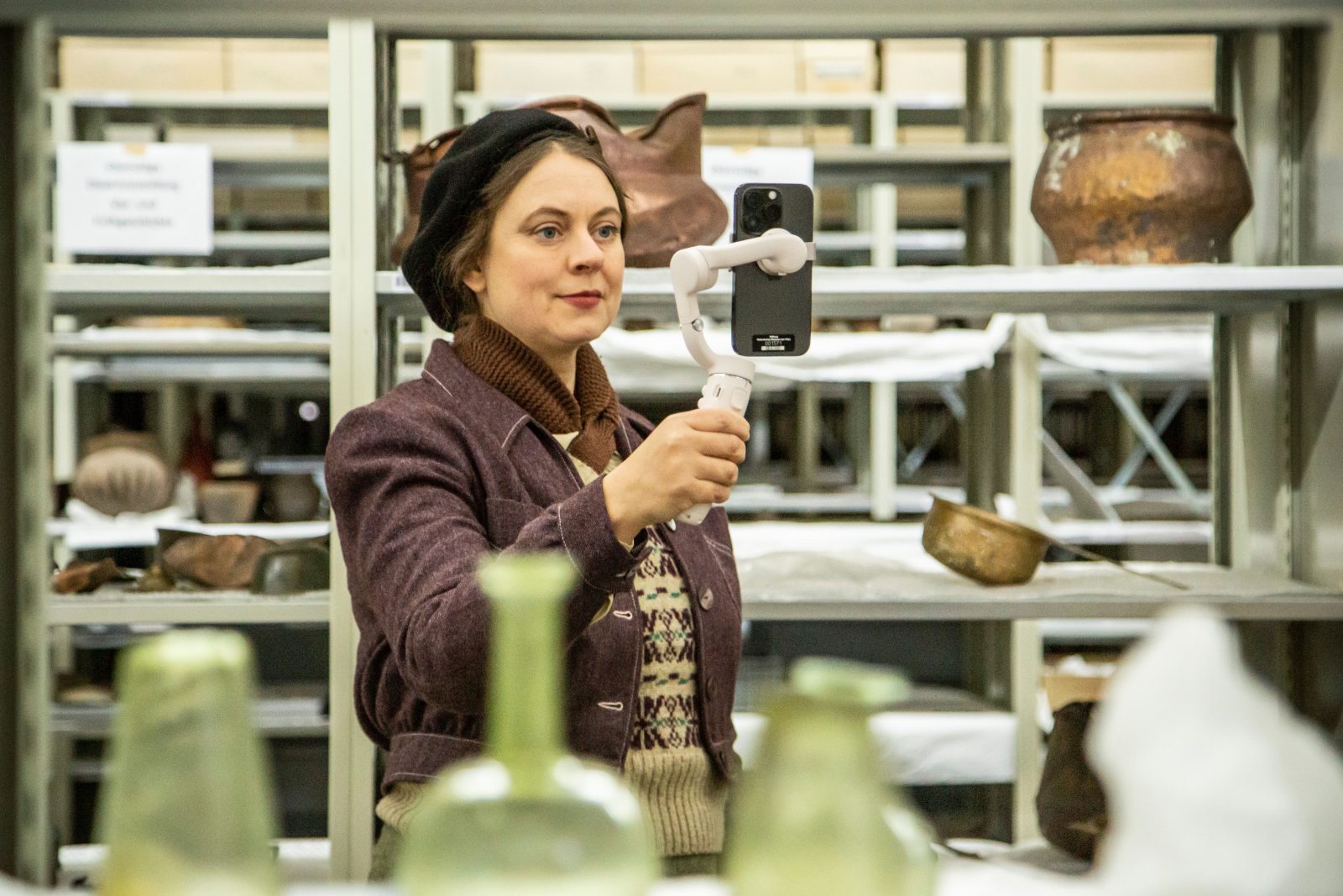 Eine Frau steht zwischen Regalen und schaut auf ein Handy. Sie filmt sich mit der Handykamera. Hinter ihr sind unterschiedliche Gegenstände aus dem Museum zu sehen.