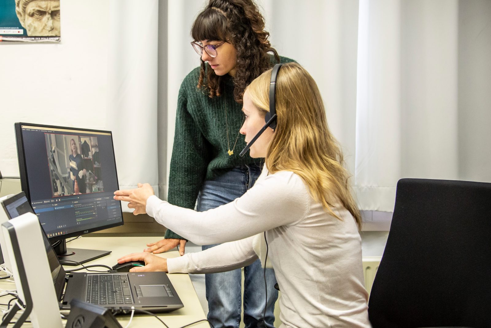 Zwei Frauen sitzen vor einem Bildschirm und einem Laptop. Sie bedienen das Regie-Progrmm für die digitalen Führungen.