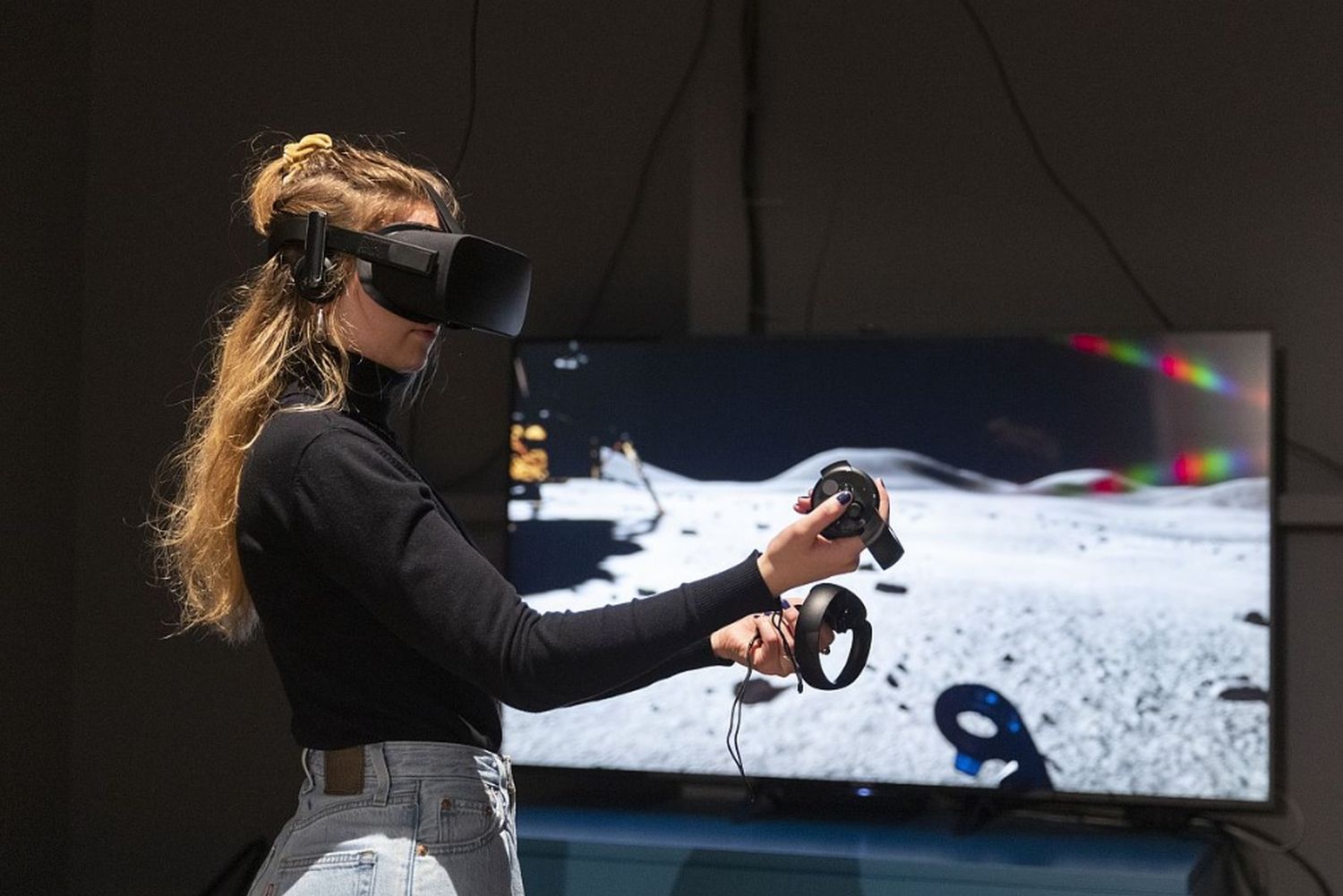 Eine Frau hat eine VR-Brille auf und hält zwei Controller in den Händen. Sie spielt eine Fahrt auf dem Mond nach.
