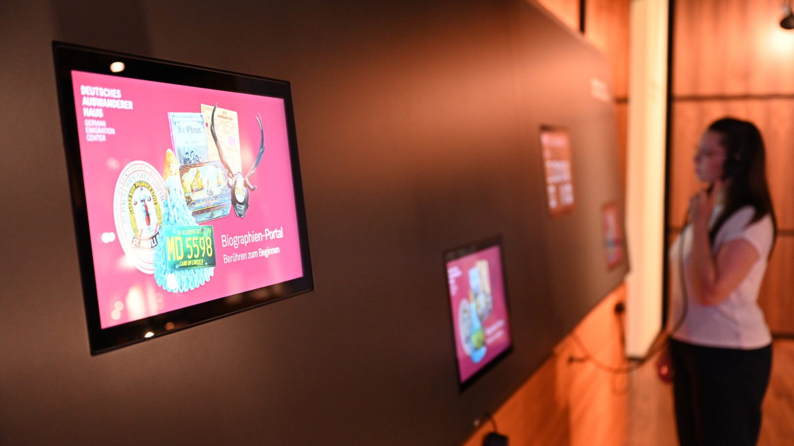 Medienstation in der musealen Ausstellung mit Objektdarstellungen. Es sind drei Screens zu sehen und eine Person, die sich einen Text daraus anhört.