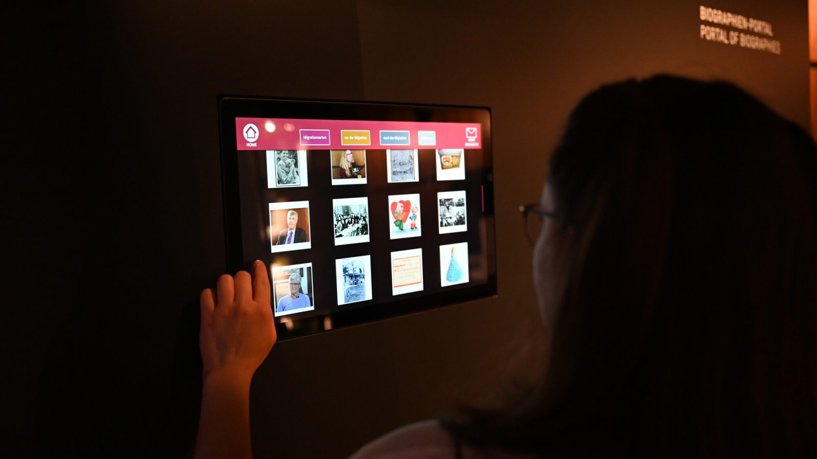 Medienstation mit Bildern von Personen und Objekten. Eine Frau steht davor und wählt auf dem Touchscreen eines der Bilder an.