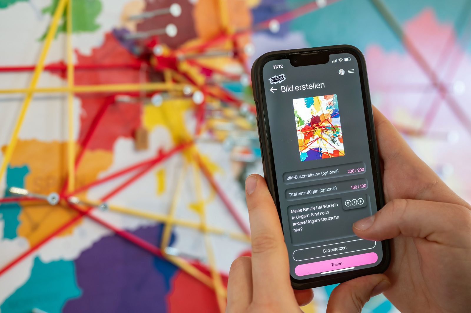 Das Bild zeigt ein Smartphone und die Hände der Nutzer*in vor einer bunten Landkarte, auf der einzelne Städte mit Schrauben markiert und mit Fäden verbunden sind. Am Smartphone ist der Screen mit Eingabefeldern zum Erstellen eines Bildbeitrags und einem Bild der Kartenwand sichtbar.