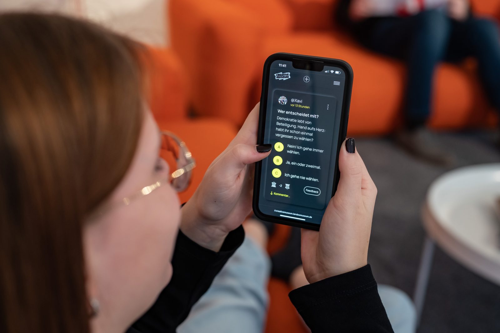 Eine junge Frau sitzt auf einem Sofa und sieht sich am Smartphone einen Beitrag im Creative Museum an.